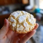 A hand holding a bright lemon cookie dusted with powdered sugar.