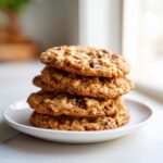 A close-up view of a stack of four soft oatmeal cookies with chocolate chips on a white plate.